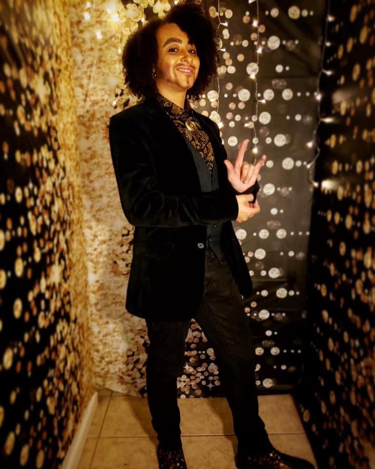 Mondo Millions, lightly melanated Black Drag King poses in front of a black and gold background. He is wearing a dark green/black velvet suit jacket with a dark grey vest, with dark jeans, and shoes with a floral pattern. He has a gold lion pendant around his neck. He is smiling and posing while holding up the one hand "I Love You" sign.